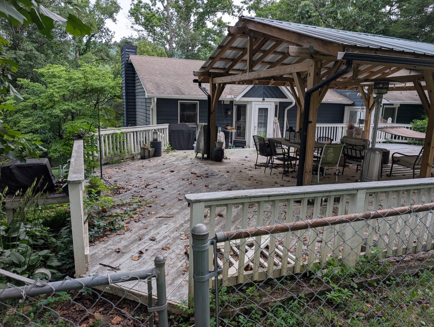 A weathered grey deck under a pavilion, leaves across the boards and vines growing through the rail, seen past a chain-link fence.