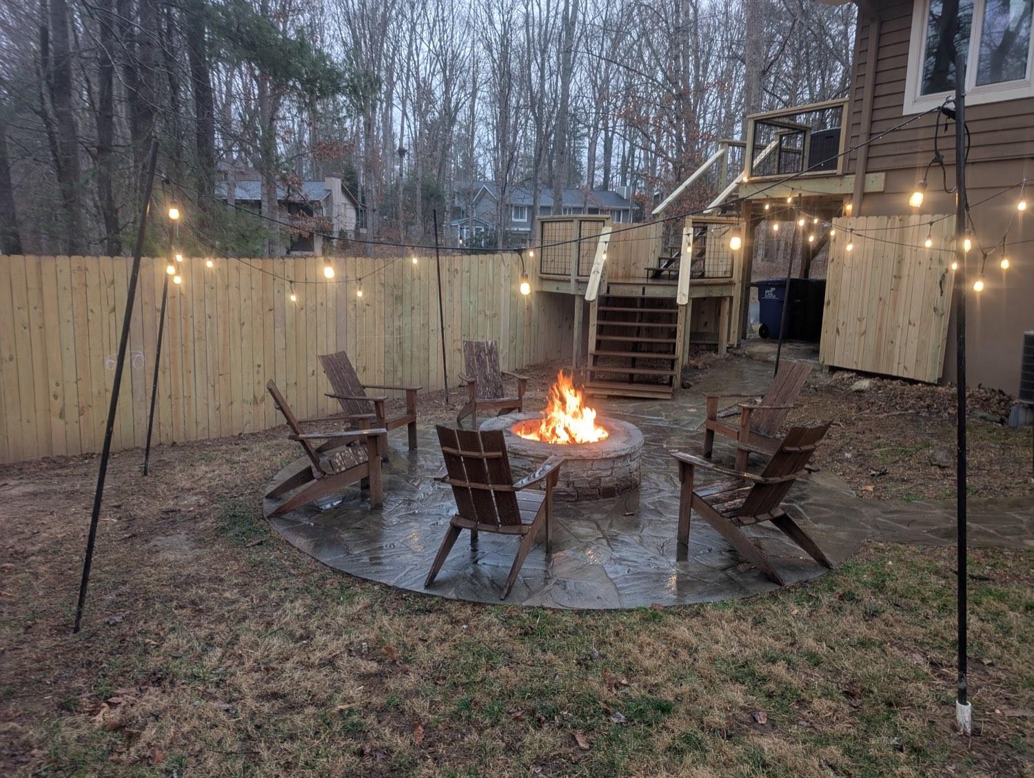 A back yard at dusk with a stone fire pit lit, string lights overhead, a new fence and a deck behind.