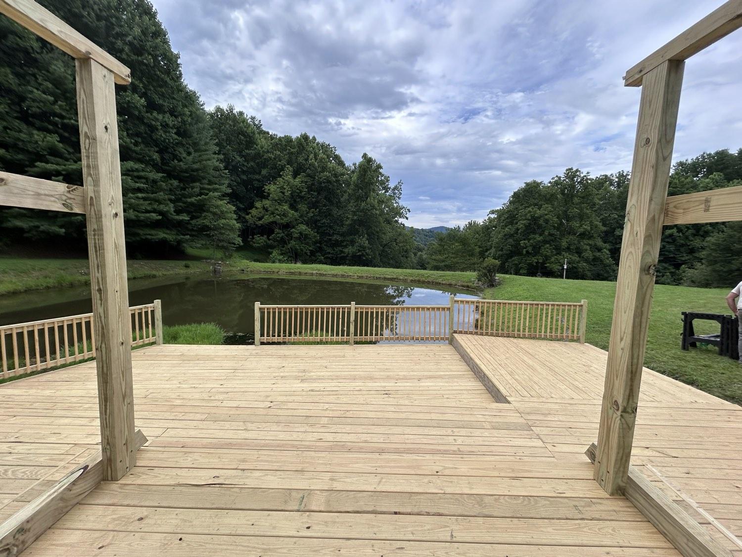 A wide pressure-treated deck framed between two pergola posts, looking across a pond to the ridges beyond.