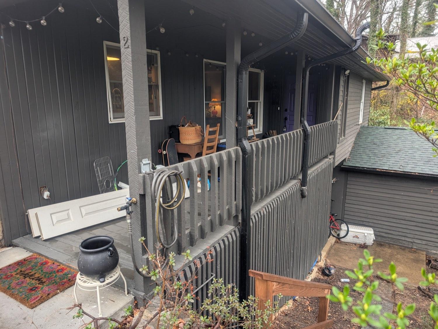 An open porch on a dark grey house with weathered pickets, a coiled hose and clutter stacked against the wall.