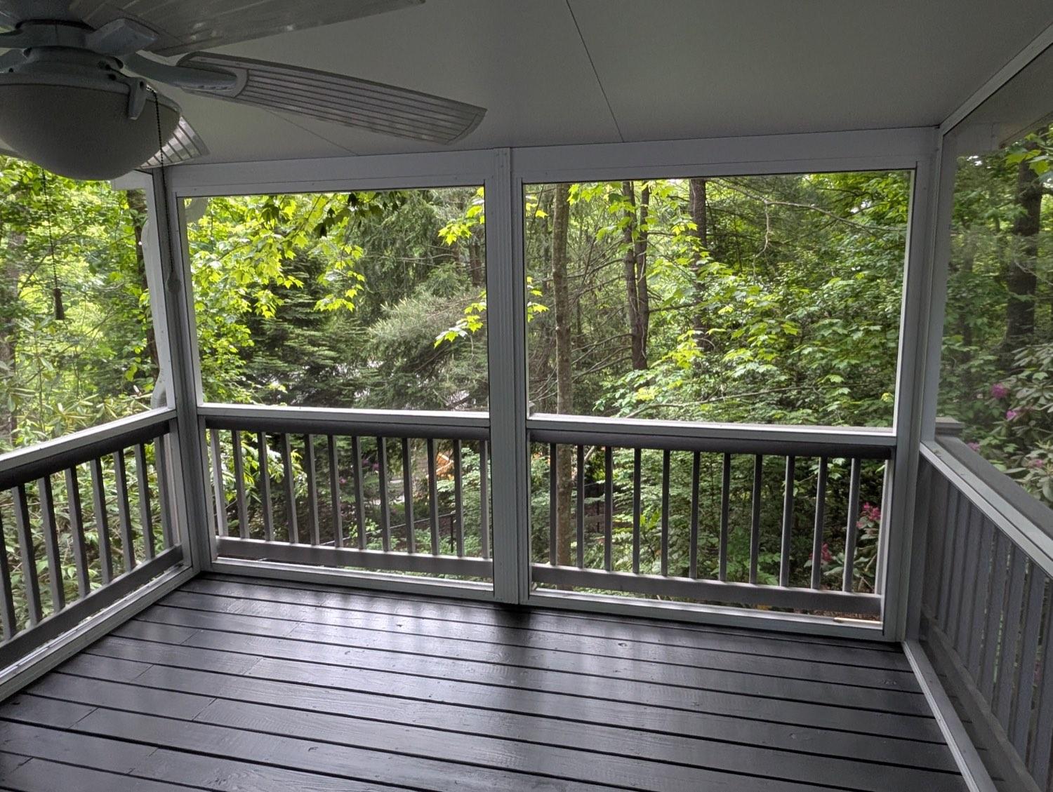 A screened porch with a ceiling fan, grey floor and full-height screens looking into green woods.