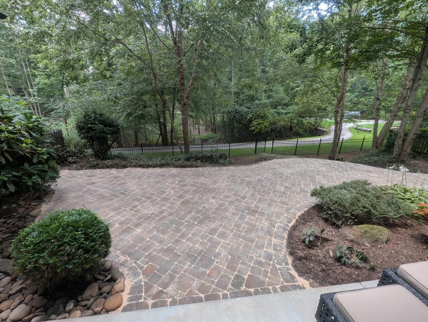 A fan-laid cobblestone patio edged with river rock and planting beds, under mature trees.