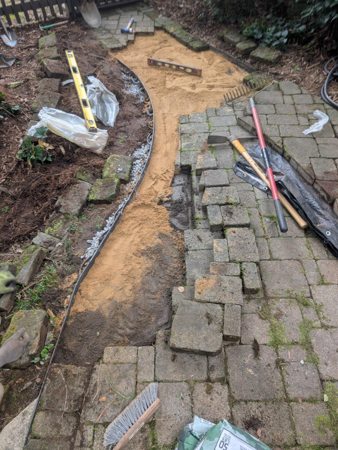 A garden path torn back to the sand base, the old cobbles stacked to one side and tools laid out along it.